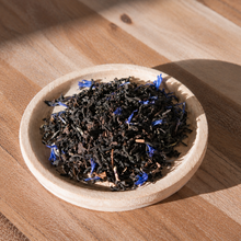 Loose leaf tea with dark black tea leaves and small blue flower petals, presented in a small round wooden dish on a wooden table.