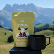 A yellow Peak Leaves 'Moo La La' tea pouch stands next to a black ceramic mug on a dark block, set against a blurred mountain landscape.