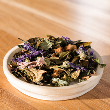 A small white ceramic dish filled with loose dried herbal tea blend, featuring green leaves, purple flowers, and brown botanical pieces, set on a warm wooden surface.