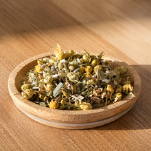 A small round wooden dish filled with dried chamomile tea ingredients, including yellow flowers and green leaves, set on a wooden table with warm sunlight and shadows.